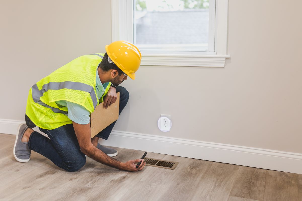 Inspector in safety vest and hard hat kneeling to examine a floor vent and flooring along a baseboard