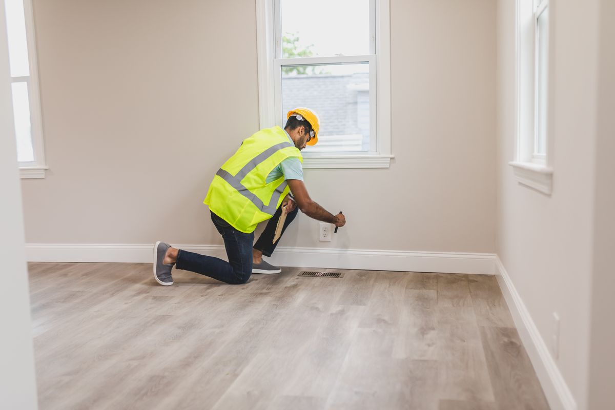 Inspector kneeling at a wall to examine an outlet and floor vent inside a home
